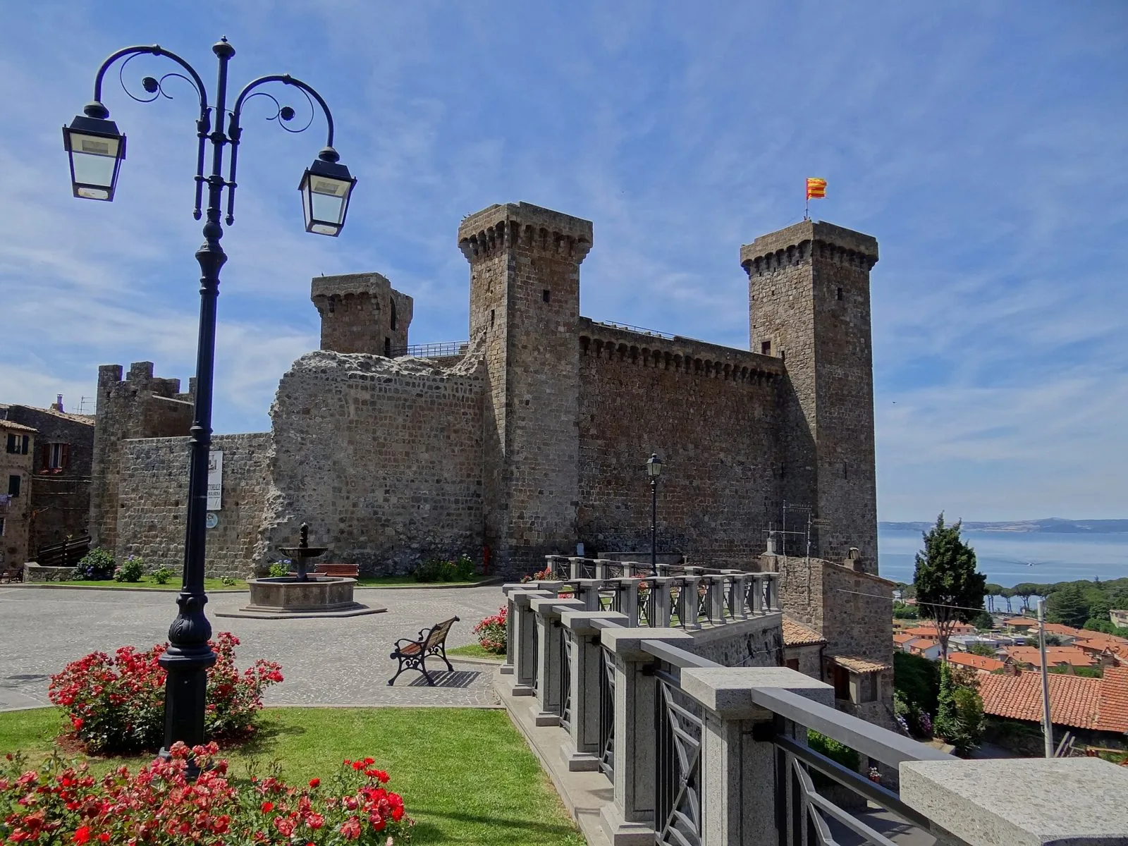 La Rocca Monaldeschi di Bolsena con le torri merlate, la piazza con fontana e lampione e il lago sullo sfondo