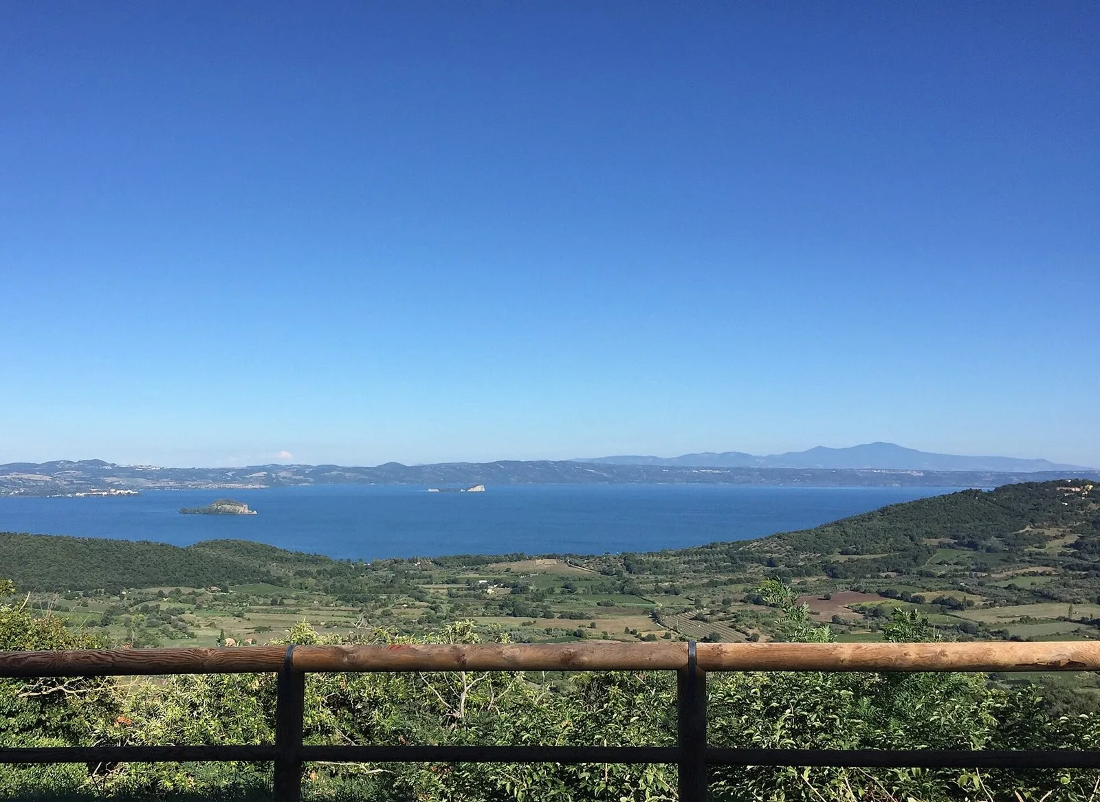 Panorama del lago di Bolsena con le isole e la campagna coltivata, visto dalla salita verso Montefiascone dietro una staccionata in legno