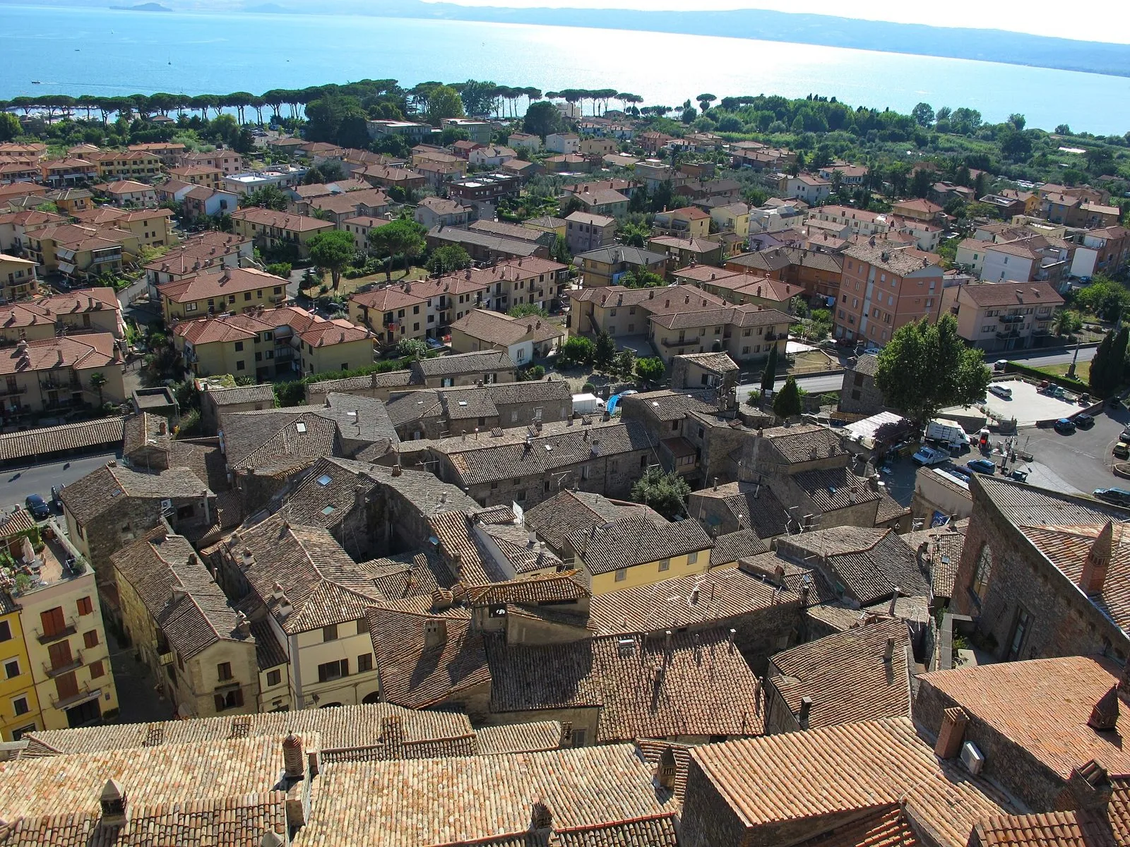 Veduta dall'alto dei tetti in coppi del centro storico di Bolsena, con la fila di pini del lungolago e il lago azzurro sullo sfondo